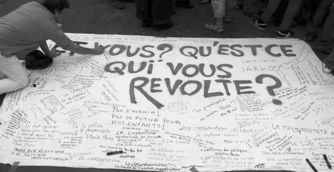 Un grand panneau blanc posé par terre avec écrit en grand: Et vous ? Qu'est-ce qui vous révolte ?
Une personne est accroupie allongée et écrit quelque chose. De nombreuses réponses sont écrites en vrac.